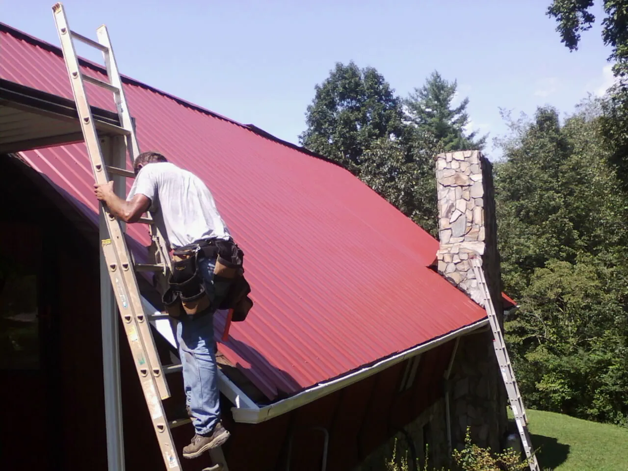 Roofer on a ladder beside a red metal roof and stone chimney — Soffit & Fascia Repair by Roofing Brothers in Mechanic Falls