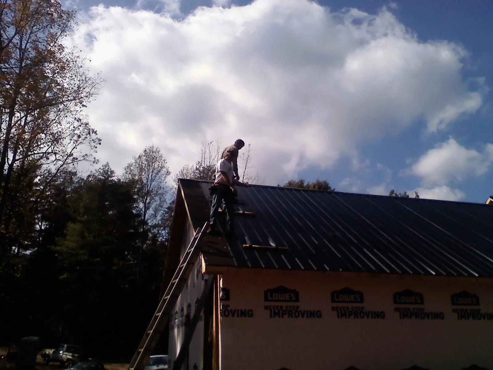 Roofer fastening dark metal panels high against a cloudy sky, from a Roof Inspection job in Mechanic Falls